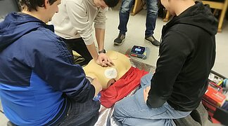 Eine Gruppe von Teilnehmern bei der praktischen Durchführung der Reanimation bestehend aus Herzdruckmassage, Beatmung sowie Anbringen und Nutzung des Defis.